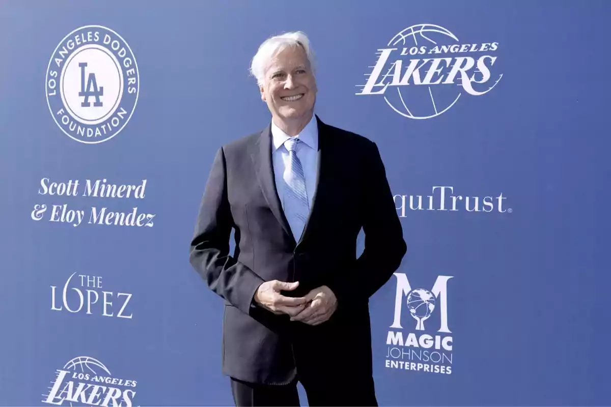 Gray-haired man in a dark suit smiles in front of a blue background with Lakers, Dodgers, and other brand logos.