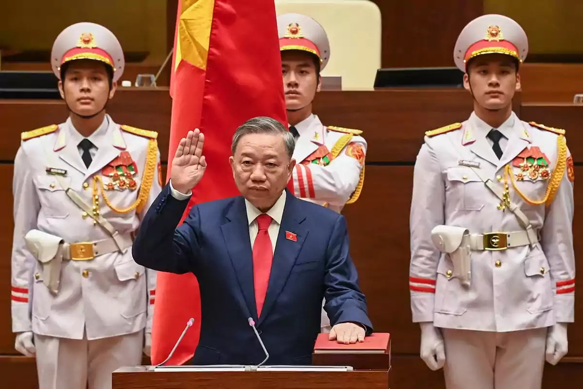 Un hombre con traje azul y corbata roja levanta la mano derecha en señal de juramento mientras tres guardias con uniforme blanco y medallas están de pie detrás de él junto a una bandera roja con una estrella amarilla