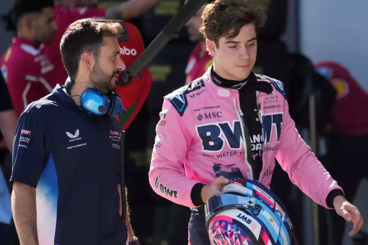 Two people in a Formula 1 paddock, one wearing a pink racing suit and holding a helmet while the other is dressed in team clothing and has headphones around their neck.