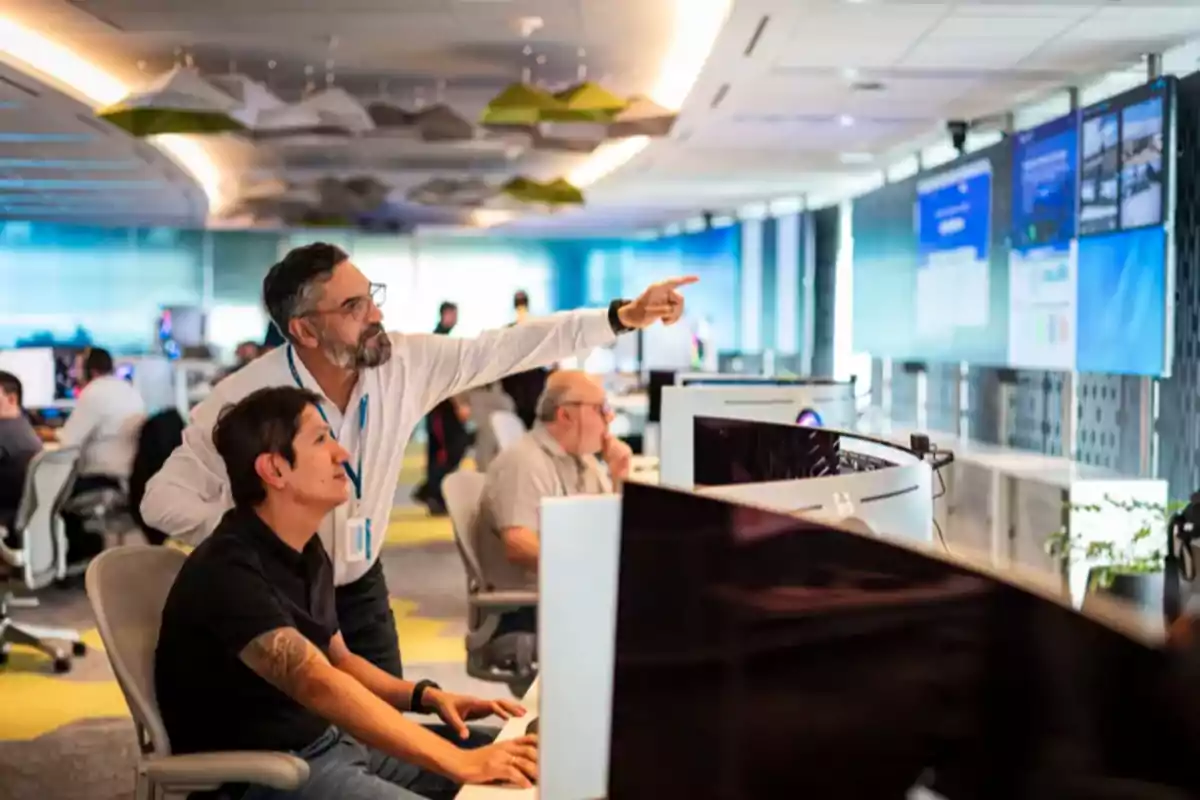 Dos personas trabajando en una oficina moderna con varias pantallas y computadoras mientras una de ellas señala hacia los monitores Dos personas trabajando en una oficina moderna con varias pantallas y computadoras mientras una de ellas señala hacia los monitores