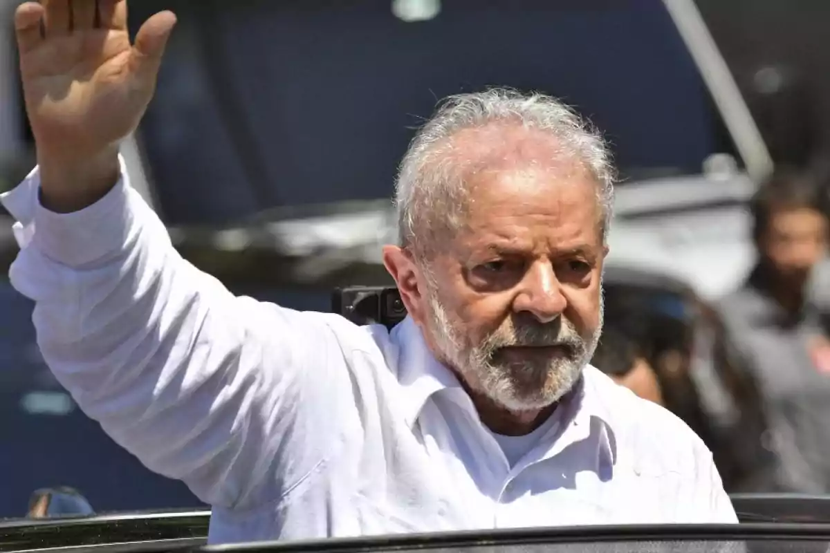 Hombre mayor con barba y cabello canoso vestido con camisa blanca levantando la mano en señal de saludo