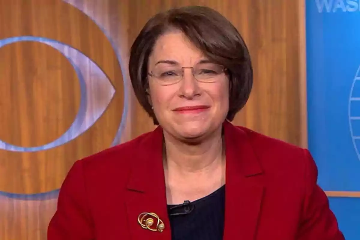 Mujer de cabello corto y gafas con saco rojo sonriendo frente a un fondo de madera y azul con logotipos de televisión Mujer de cabello corto y gafas con saco rojo sonriendo frente a un fondo de madera y azul con logotipos de televisión