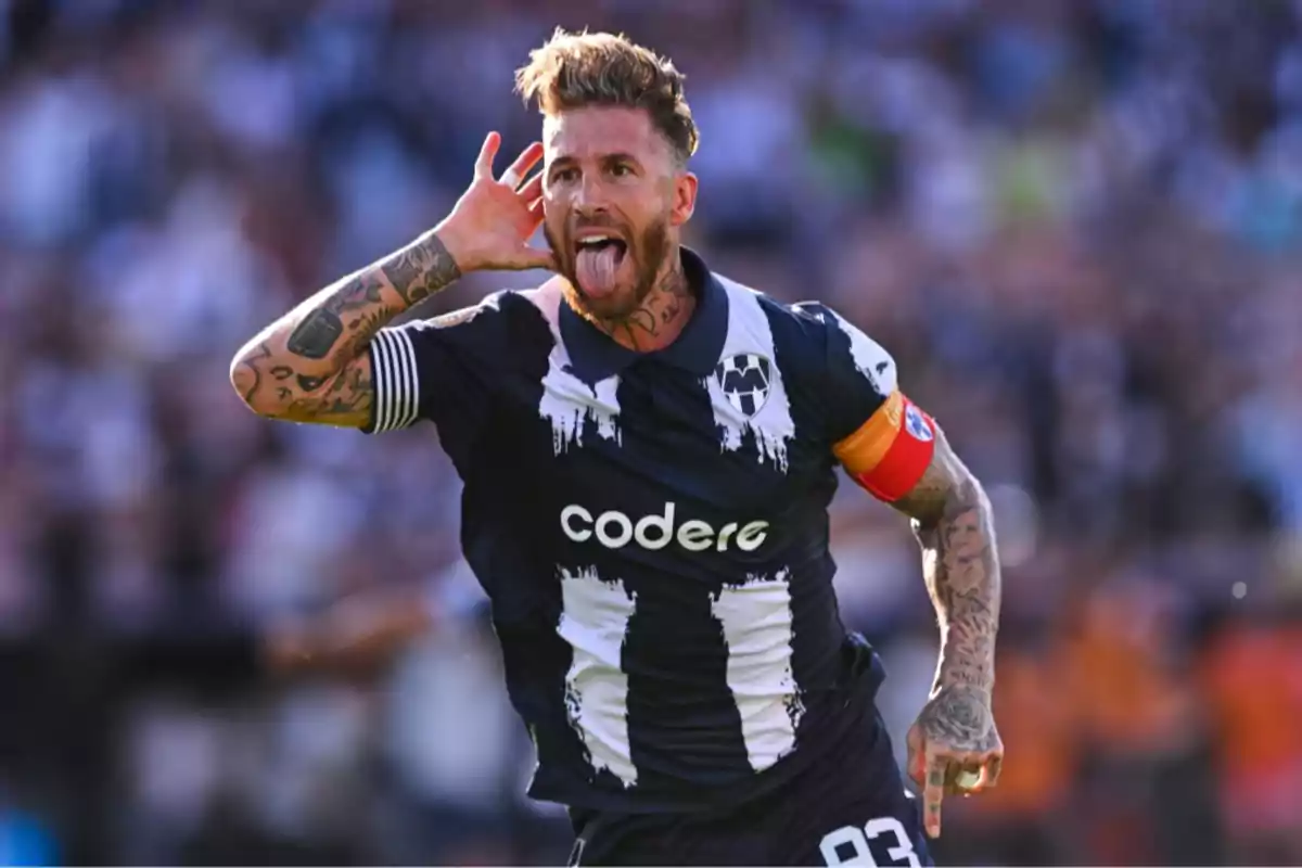 Futbolista celebrando un gol con la camiseta de Rayados de Monterrey llevando el gafete de capitán y haciendo un gesto con la mano en la oreja y la lengua afuera Futbolista celebrando un gol con la camiseta de Rayados de Monterrey llevando el gafete de capitán y haciendo un gesto con la mano en la oreja y la lengua afuera