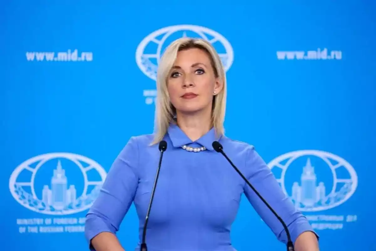 Blonde woman in a blue dress speaking in front of two microphones with a blue background and official logos Blonde woman in a blue dress speaking in front of two microphones with a blue background and official logos