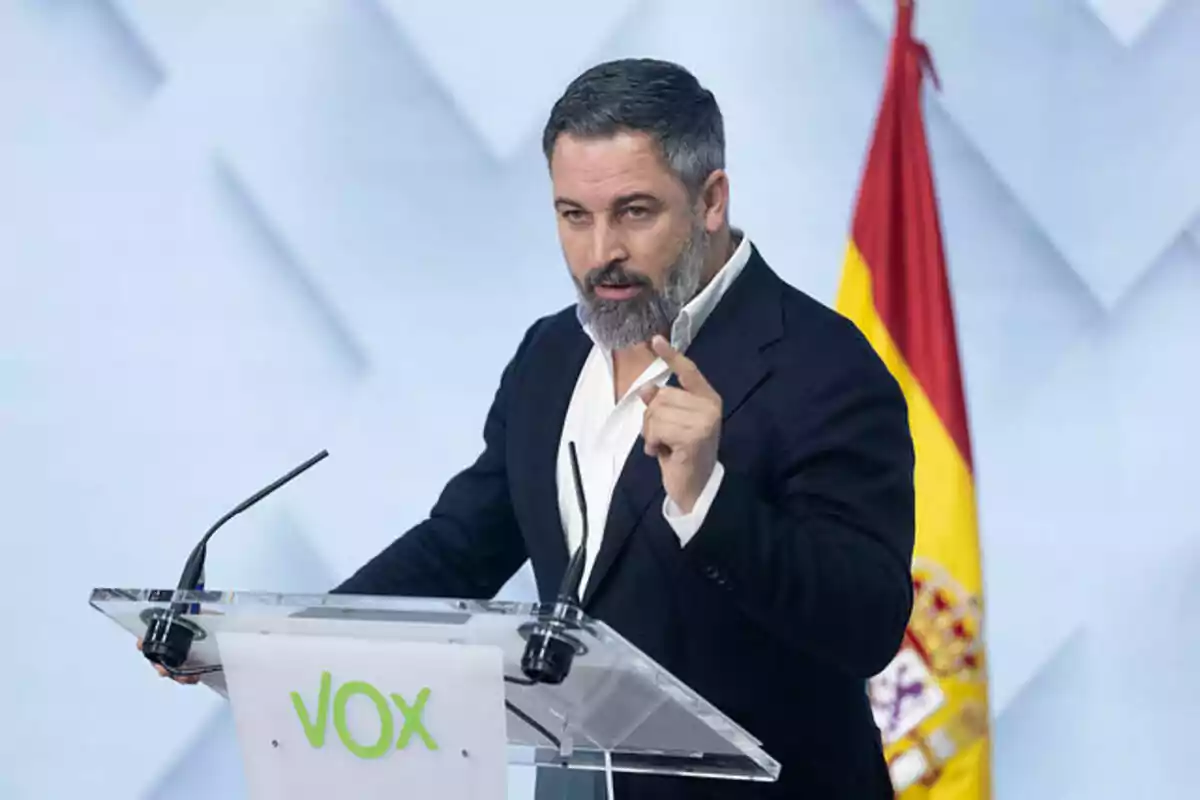 Hombre de barba y cabello corto hablando en un atril con el logo de VOX y una bandera de España al fondo Hombre de barba y cabello corto hablando en un atril con el logo de VOX y una bandera de España al fondo