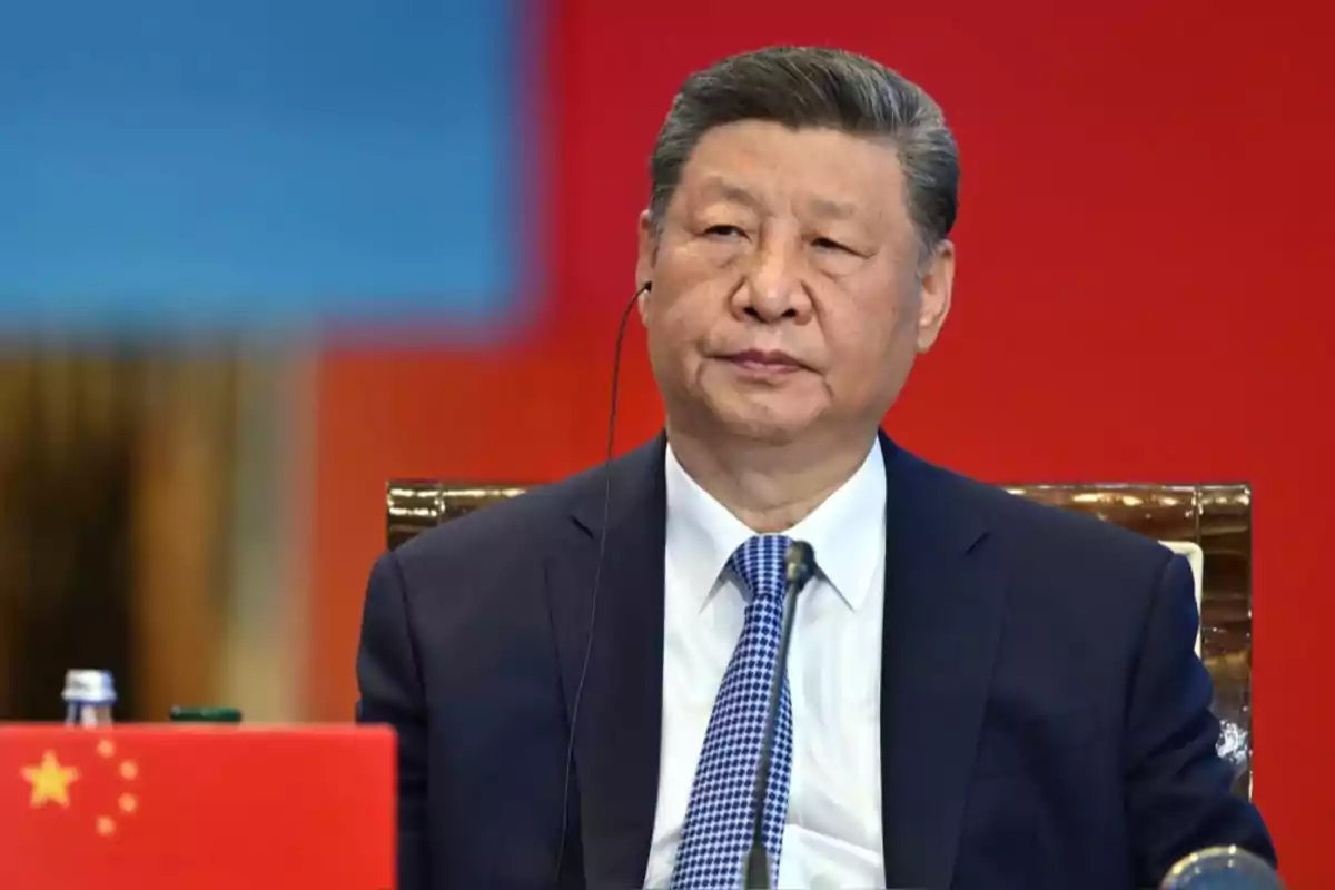 Man in a dark suit and blue tie sitting in front of a red and blue background with a Chinese flag in the foreground Man in a dark suit and blue tie sitting in front of a red and blue background with a Chinese flag in the foreground
