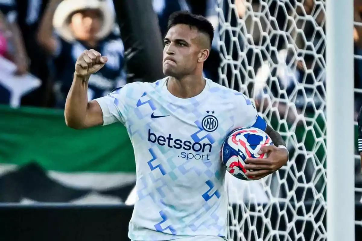 Futbolista con uniforme blanco celebrando con el puño en alto mientras sostiene un balón frente a la portería Futbolista con uniforme blanco celebrando con el puño en alto mientras sostiene un balón frente a la portería