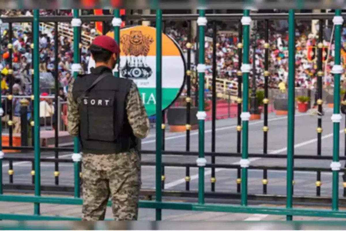 Un guardia con uniforme militar y boina roja está de espaldas frente a una reja, al fondo se observa una multitud y un letrero con los colores de la bandera de la India. Un guardia con uniforme militar y boina roja está de espaldas frente a una reja, al fondo se observa una multitud y un letrero con los colores de la bandera de la India.