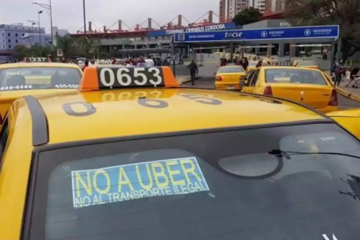 Several yellow taxis parked in front of a bus terminal with a sign in the rear window of one of them that says NO TO UBER NO TO ILLEGAL TRANSPORTATION Several yellow taxis parked in front of a bus terminal with a sign in the rear window of one of them that says NO TO UBER NO TO ILLEGAL TRANSPORTATION