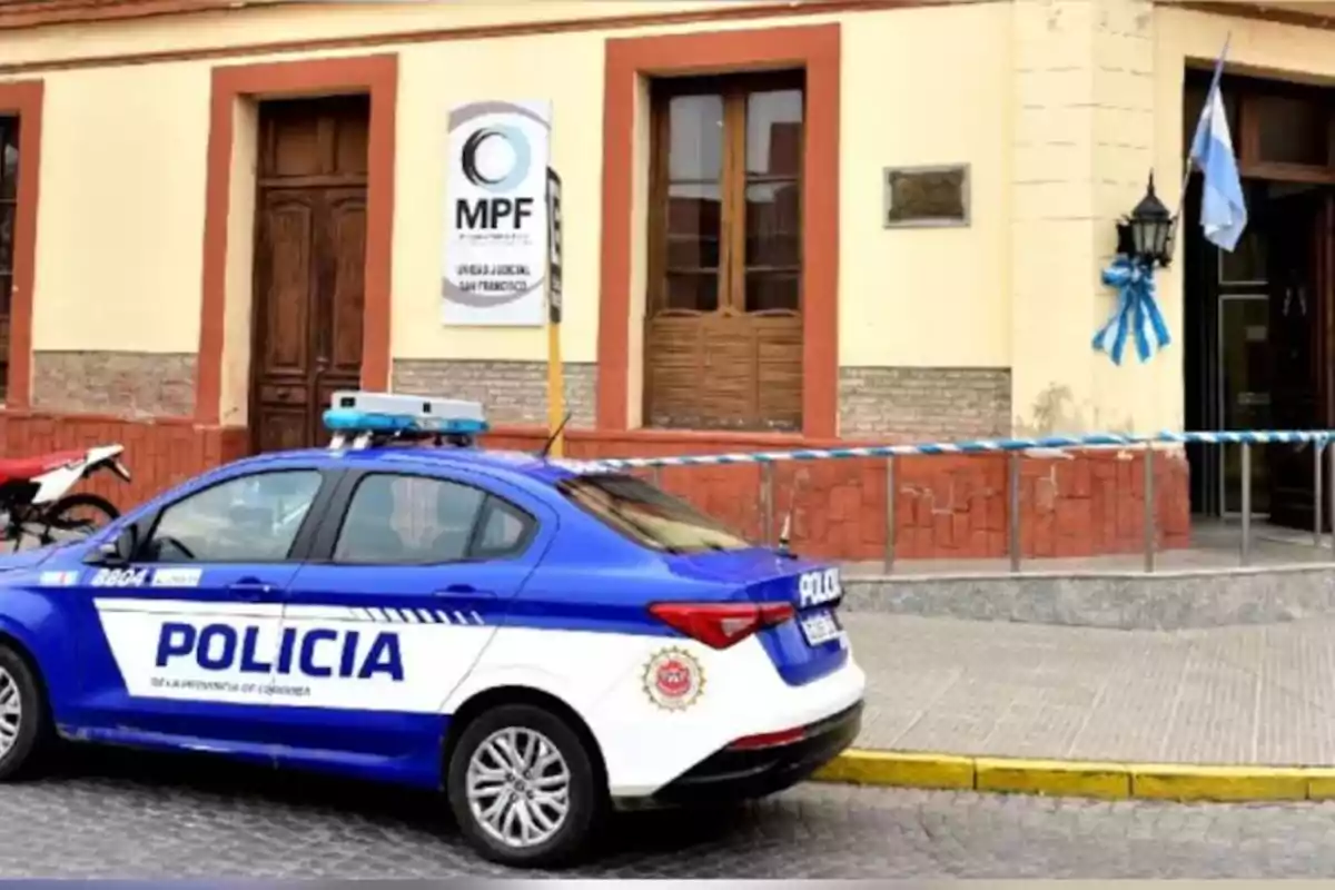 Un patrullero de la policía de Córdoba estacionado frente a un edificio público con una bandera argentina y cintas de seguridad Un patrullero de la policía de Córdoba estacionado frente a un edificio público con una bandera argentina y cintas de seguridad