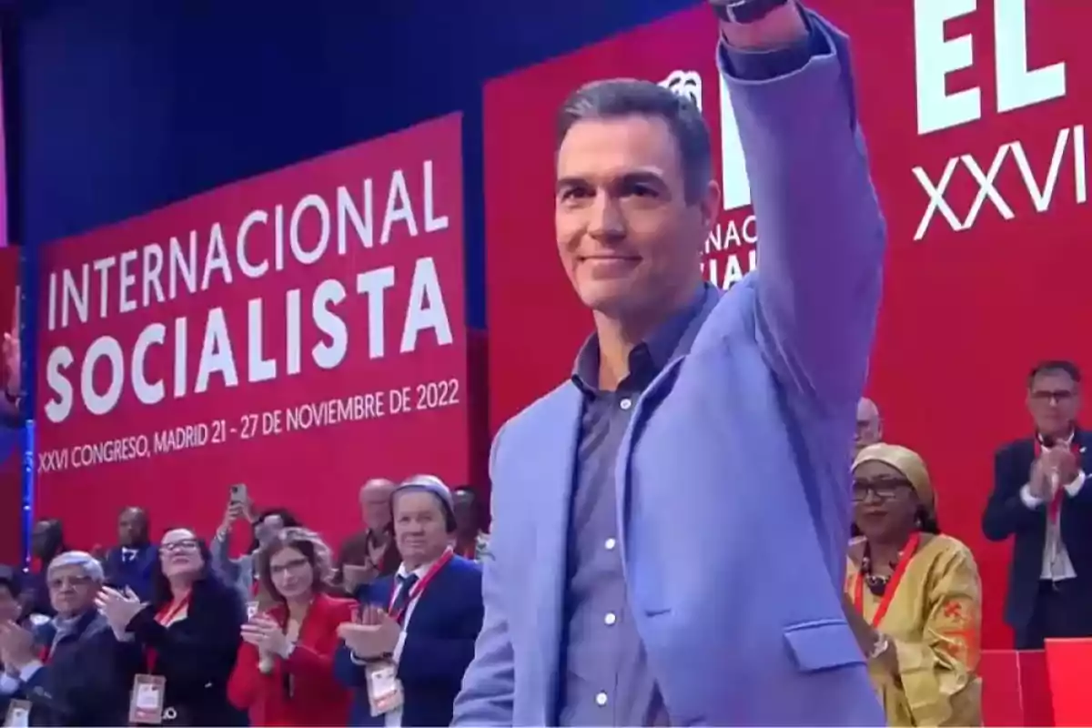 Hombre con chaqueta azul saluda en un evento del Congreso Internacional Socialista en Madrid mientras varias personas aplauden en el fondo Hombre con chaqueta azul saluda en un evento del Congreso Internacional Socialista en Madrid mientras varias personas aplauden en el fondo