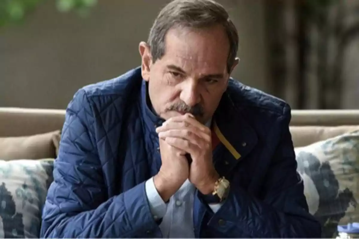 Hombre de mediana edad con bigote y chaqueta azul sentado en un sofá con las manos entrelazadas y expresión pensativa
