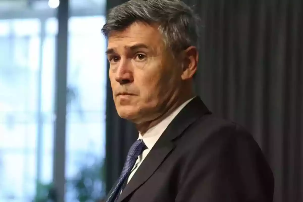 Hombre de cabello canoso con traje oscuro y corbata azul mirando hacia un lado en un entorno interior con cortinas oscuras de fondo Hombre de cabello canoso con traje oscuro y corbata azul mirando hacia un lado en un entorno interior con cortinas oscuras de fondo