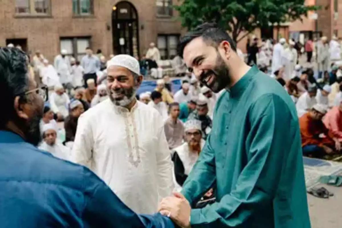 Three men smile and shake hands at an outdoor gathering with many people in the background, all dressed in traditional clothing. Three men smile and shake hands at an outdoor gathering with many people in the background, all dressed in traditional clothing.
