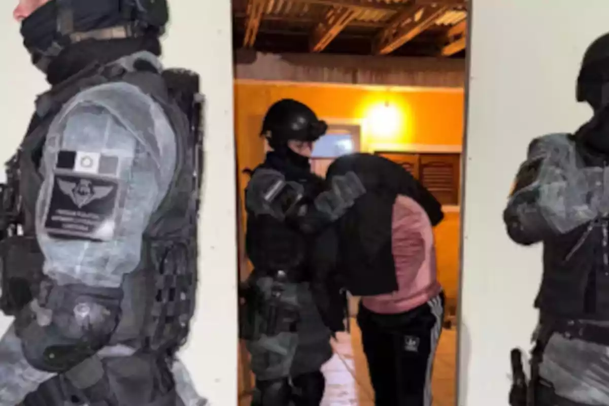Elementos policiales fuertemente armados detienen a una persona que cubre su rostro con una prenda dentro de una vivienda iluminada. Elementos policiales fuertemente armados detienen a una persona que cubre su rostro con una prenda dentro de una vivienda iluminada.