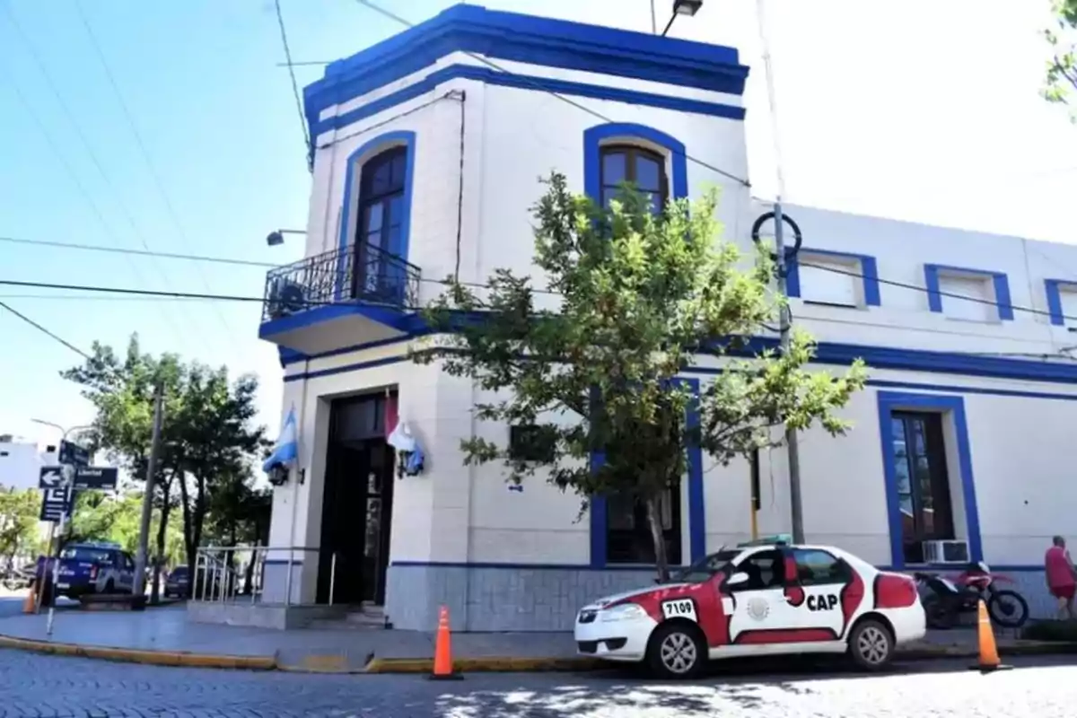 Edificio de dos pisos con detalles azules en la esquina de una calle, un patrullero estacionado al frente y árboles alrededor Edificio de dos pisos con detalles azules en la esquina de una calle, un patrullero estacionado al frente y árboles alrededor