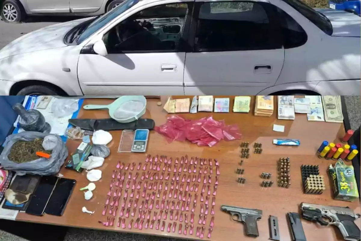 Un coche blanco estacionado junto a una mesa que exhibe armas, municiones, dinero en efectivo, drogas, teléfonos móviles, bolsas plásticas y utensilios diversos.