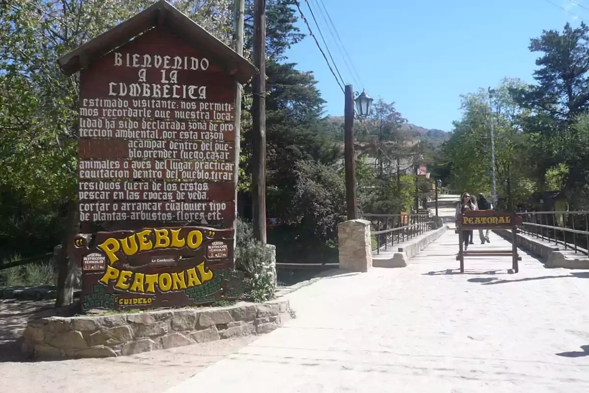 Cartel de bienvenida de madera en la entrada de un pueblo peatonal rodeado de árboles y un puente donde caminan algunas personas Cartel de bienvenida de madera en la entrada de un pueblo peatonal rodeado de árboles y un puente donde caminan algunas personas