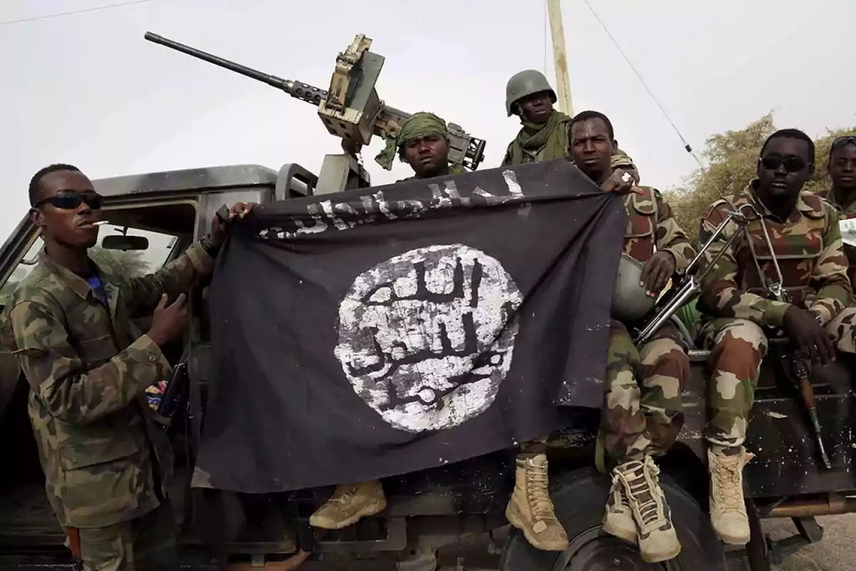A group of soldiers in camouflage uniforms holds a black flag with Arabic inscriptions while sitting on an armed military vehicle. A group of soldiers in camouflage uniforms holds a black flag with Arabic inscriptions while sitting on an armed military vehicle.