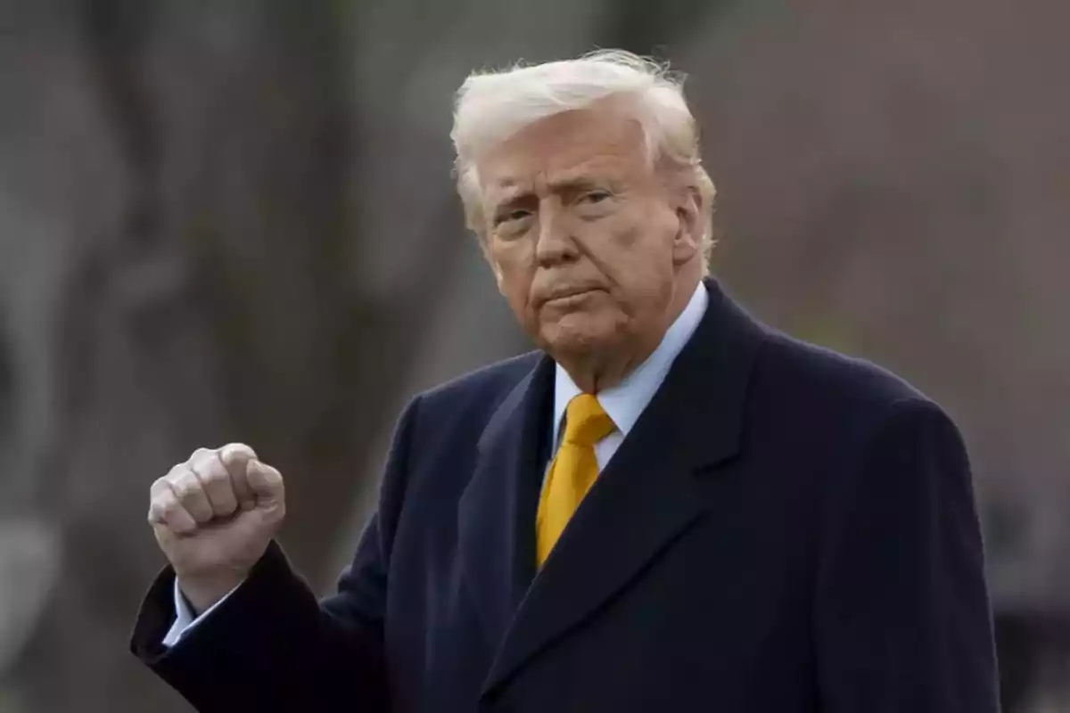 Hombre de cabello rubio con abrigo oscuro y corbata amarilla levantando el puño