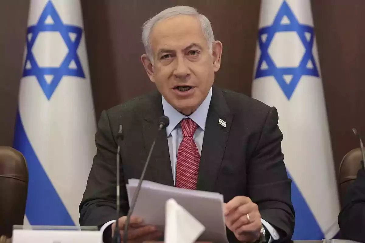 Hombre de cabello canoso con traje oscuro y corbata roja hablando frente a micrófonos con banderas de Israel al fondo Hombre de cabello canoso con traje oscuro y corbata roja hablando frente a micrófonos con banderas de Israel al fondo