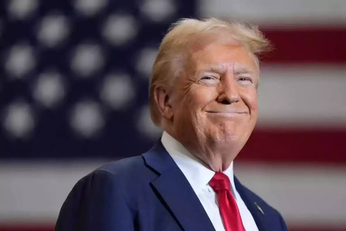 Blond-haired man in a blue suit and red tie smiling in front of a blurred United States flag Blond-haired man in a blue suit and red tie smiling in front of a blurred United States flag