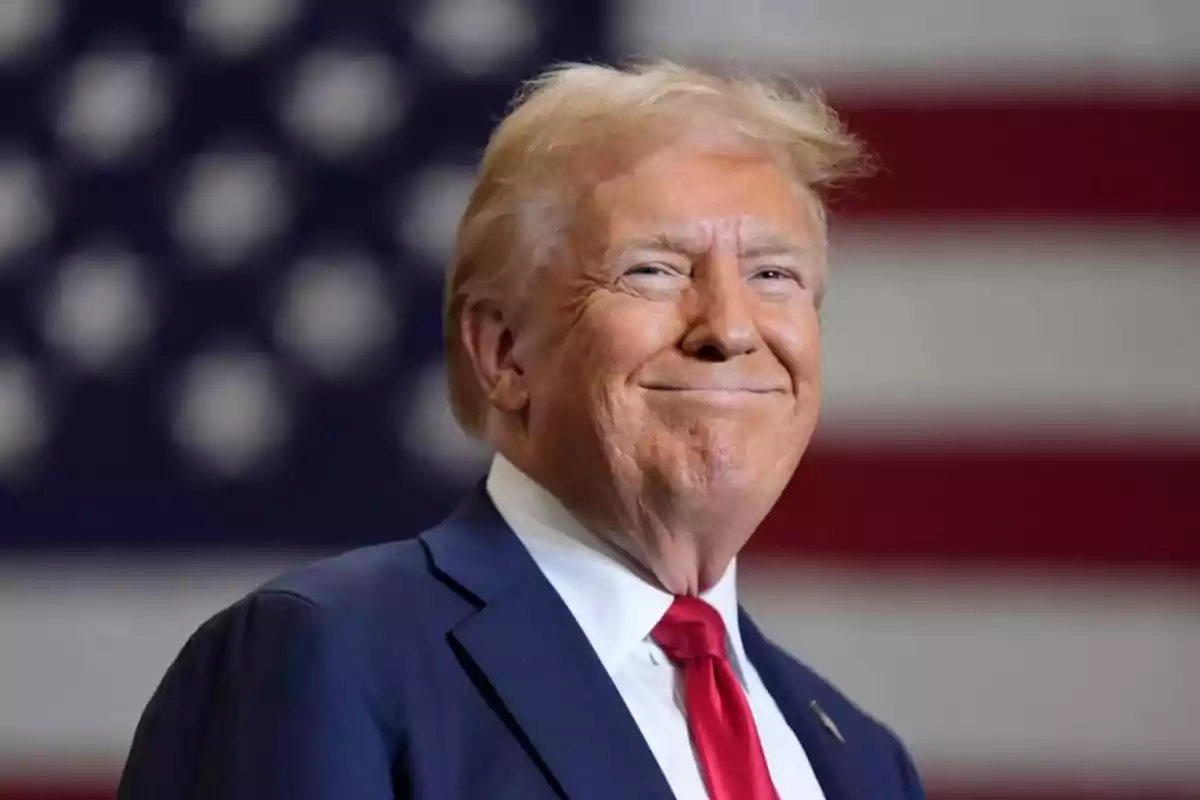 Hombre de cabello rubio con traje azul y corbata roja sonriendo frente a una bandera de Estados Unidos desenfocada