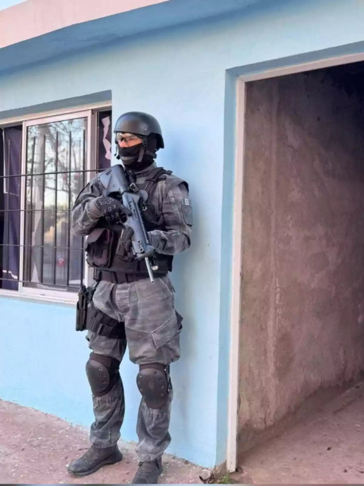 Policía fuertemente armado con uniforme táctico y casco de protección parado junto a la entrada de una casa de paredes azules Policía fuertemente armado con uniforme táctico y casco de protección parado junto a la entrada de una casa de paredes azules