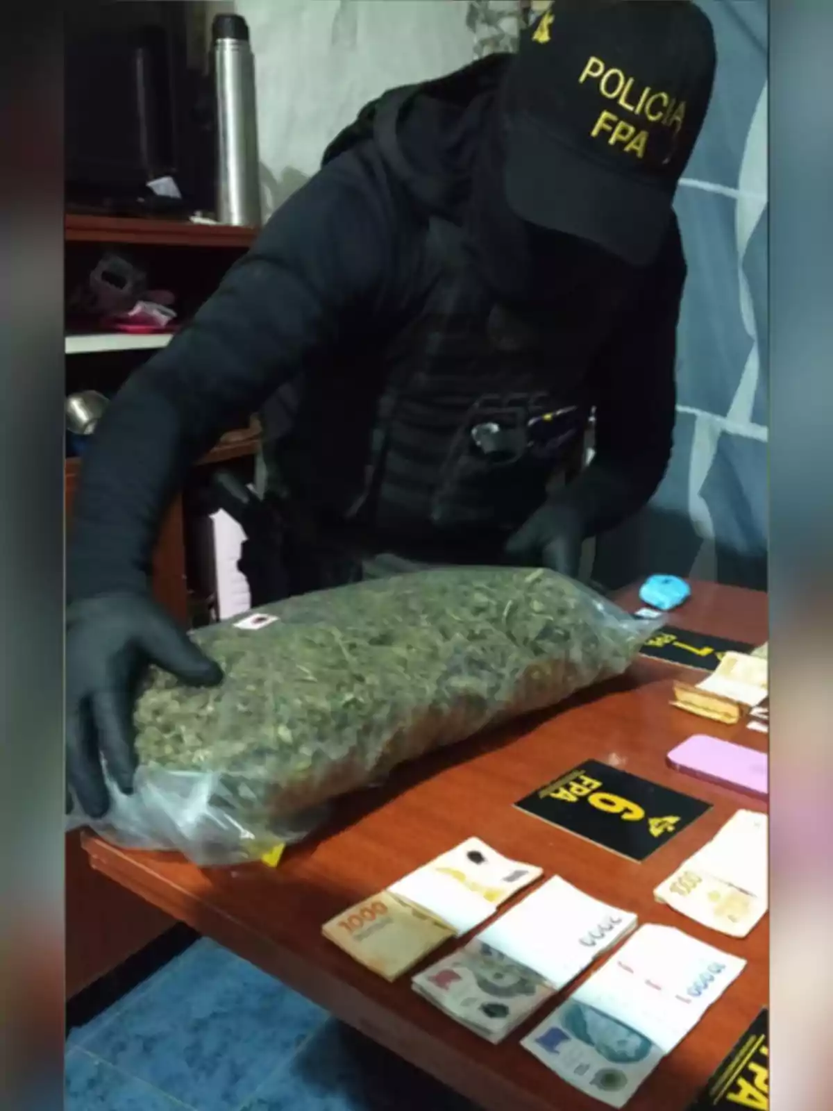 Agente policial encapuchado con uniforme de la FPA manipulando una bolsa grande de marihuana sobre una mesa donde también hay billetes y elementos incautados Agente policial encapuchado con uniforme de la FPA manipulando una bolsa grande de marihuana sobre una mesa donde también hay billetes y elementos incautados