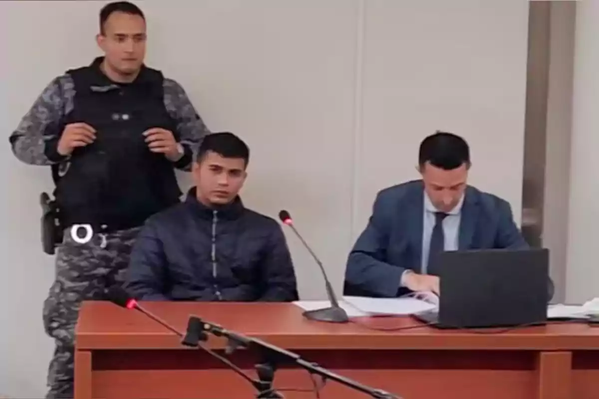 A man in a dark jacket is sitting next to a lawyer who is reviewing documents on a laptop while a standing security guard watches them in a courtroom. A man in a dark jacket is sitting next to a lawyer who is reviewing documents on a laptop while a standing security guard watches them in a courtroom.