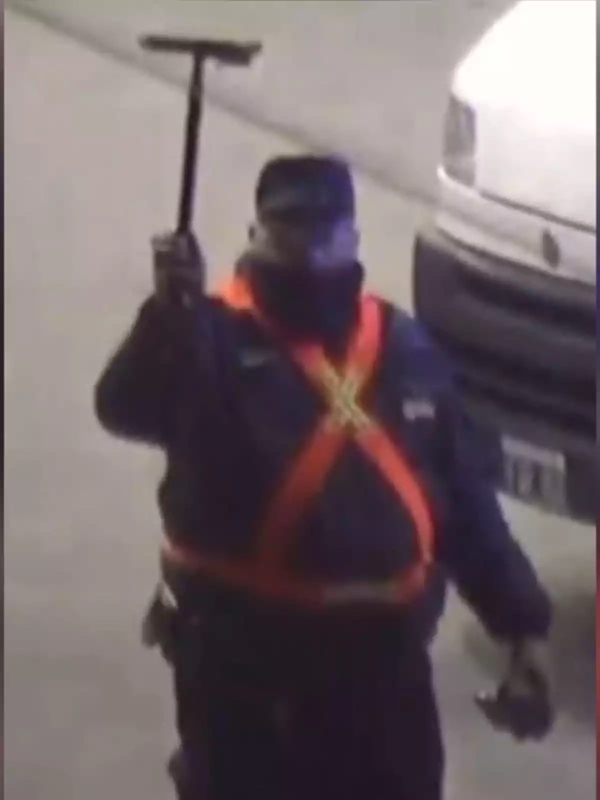 Persona con uniforme oscuro y chaleco reflectante naranja sosteniendo una herramienta en alto frente a un vehículo blanco Persona con uniforme oscuro y chaleco reflectante naranja sosteniendo una herramienta en alto frente a un vehículo blanco