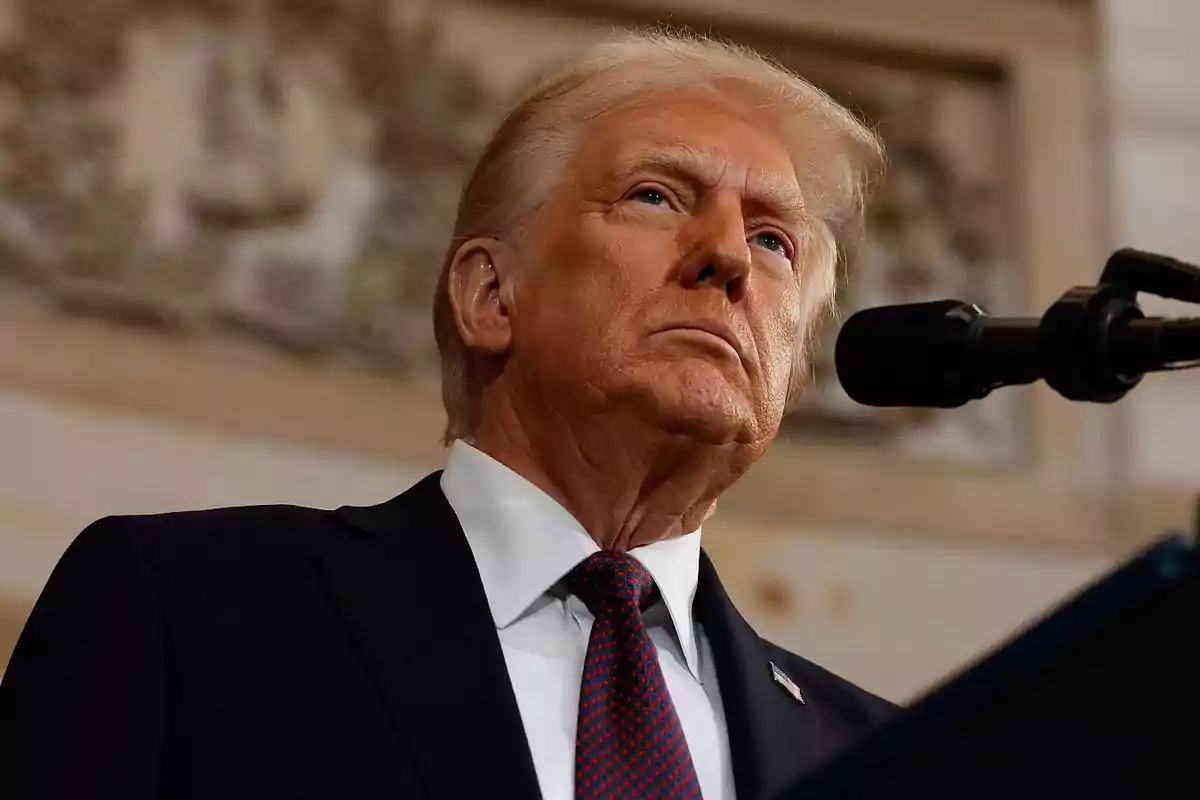 Hombre de cabello rubio con traje oscuro y corbata roja hablando frente a un micrófono en un entorno formal Hombre de cabello rubio con traje oscuro y corbata roja hablando frente a un micrófono en un entorno formal