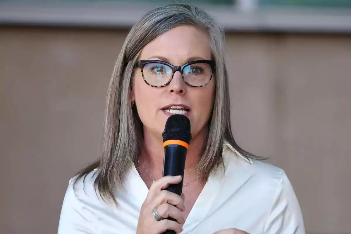 Mujer de cabello canoso y gafas hablando mientras sostiene un micrófono Mujer de cabello canoso y gafas hablando mientras sostiene un micrófono