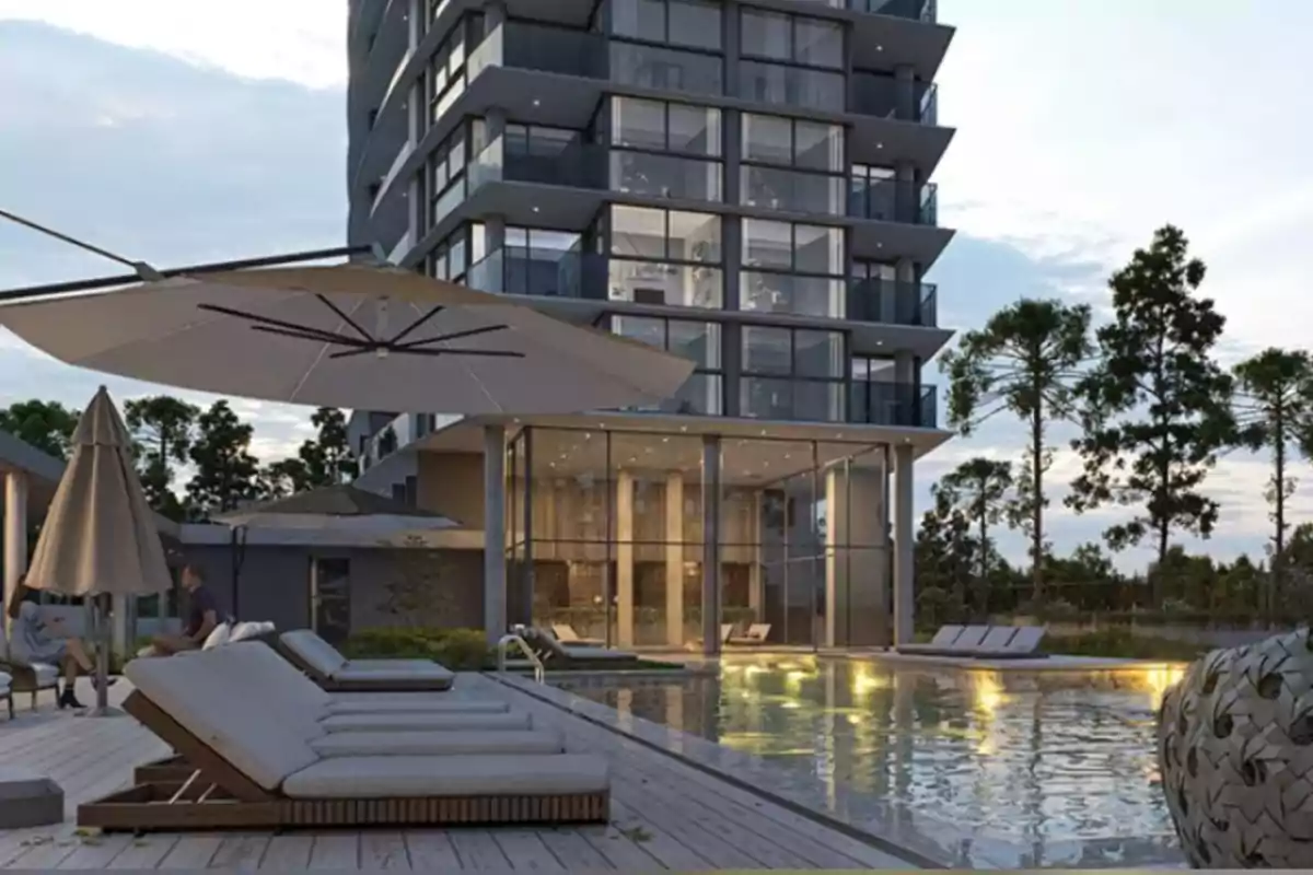 Edificio moderno de varios pisos con grandes ventanales y balcones, área de piscina iluminada, camastros y sombrillas rodeados de árboles y vegetación Edificio moderno de varios pisos con grandes ventanales y balcones, área de piscina iluminada, camastros y sombrillas rodeados de árboles y vegetación