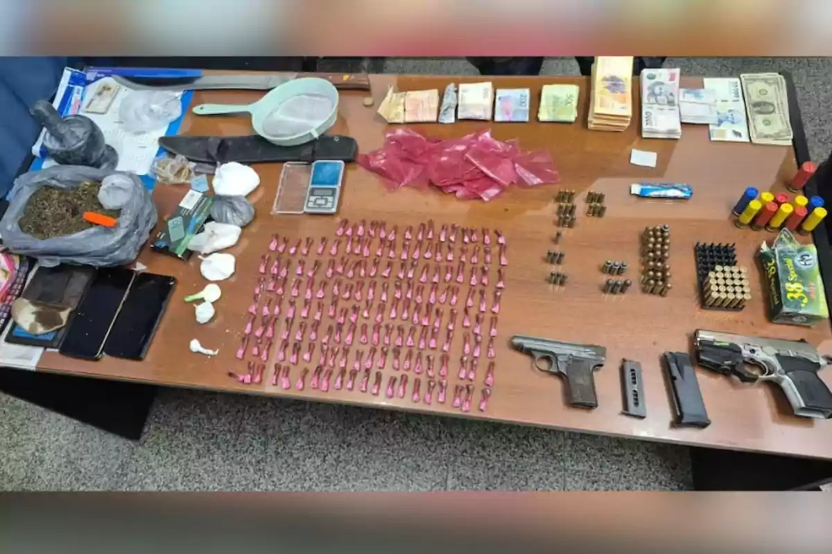 Mesa con armas, municiones, dinero en efectivo, teléfonos móviles, bolsas con sustancias, una balanza, cuchillos, cartuchos y otros objetos relacionados con actividades ilícitas. Mesa con armas, municiones, dinero en efectivo, teléfonos móviles, bolsas con sustancias, una balanza, cuchillos, cartuchos y otros objetos relacionados con actividades ilícitas.