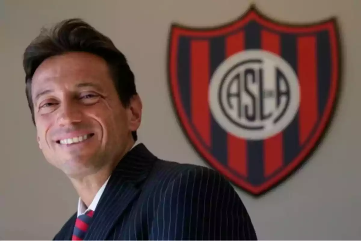Smiling man in a suit in front of the San Lorenzo de Almagro club crest