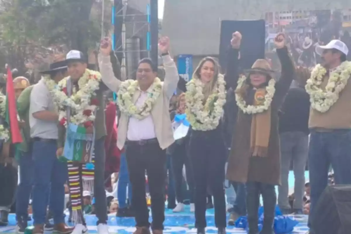 Grupo de personas en un escenario al aire libre con guirnaldas de flores alrededor del cuello y los brazos levantados en señal de celebración Grupo de personas en un escenario al aire libre con guirnaldas de flores alrededor del cuello y los brazos levantados en señal de celebración