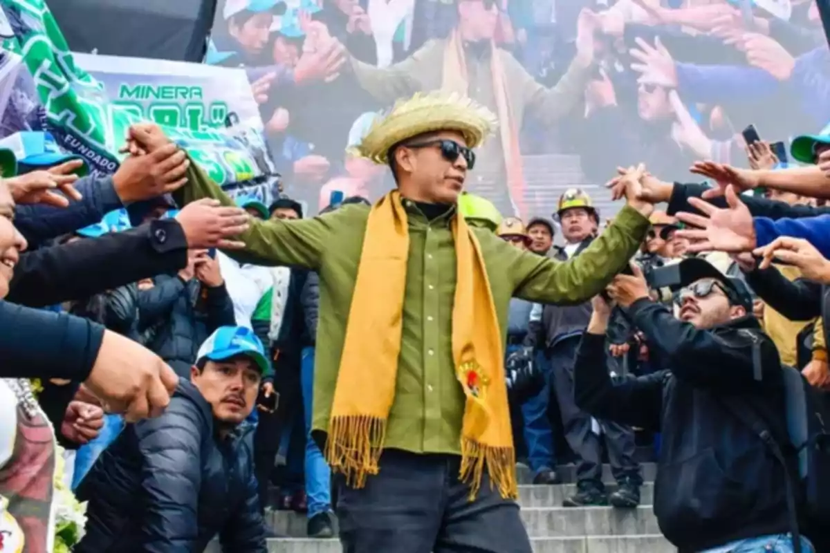 Hombre con sombrero de paja y bufanda amarilla es recibido por una multitud que le extiende las manos mientras camina por unas escaleras Hombre con sombrero de paja y bufanda amarilla es recibido por una multitud que le extiende las manos mientras camina por unas escaleras