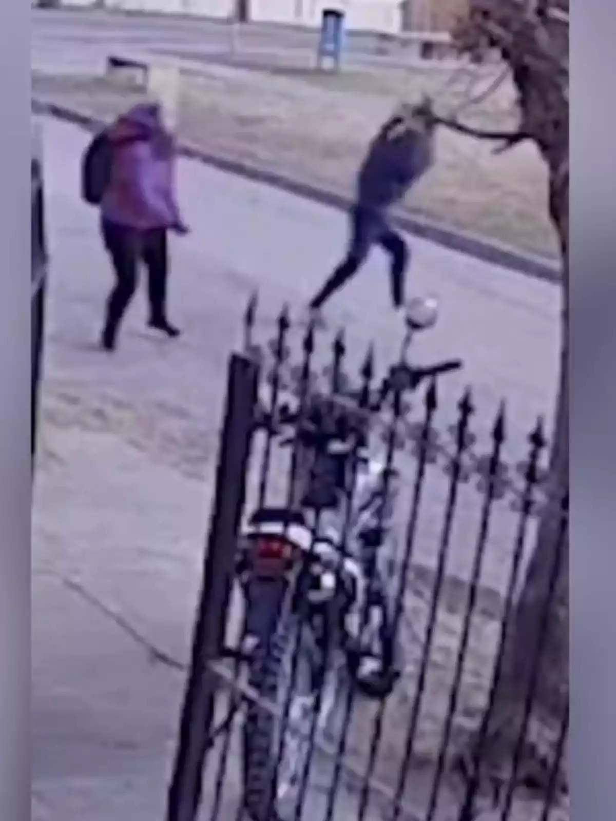 Dos personas caminando por una acera mientras una motocicleta está estacionada detrás de una reja en primer plano Dos personas caminando por una acera mientras una motocicleta está estacionada detrás de una reja en primer plano