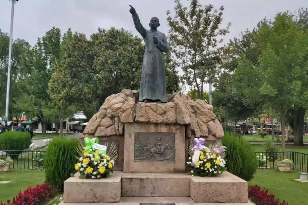Estatua de un hombre con vestimenta religiosa señalando hacia el cielo sobre una base de piedra rodeada de arreglos florales en un parque con árboles y césped Estatua de un hombre con vestimenta religiosa señalando hacia el cielo sobre una base de piedra rodeada de arreglos florales en un parque con árboles y césped