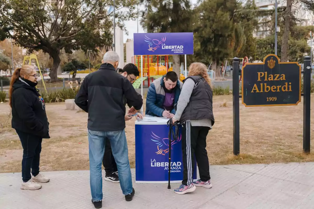 Varias personas reunidas alrededor de una mesa de un partido político en una plaza pública junto a un cartel que indica Plaza Alberdi 1909 Varias personas reunidas alrededor de una mesa de un partido político en una plaza pública junto a un cartel que indica Plaza Alberdi 1909