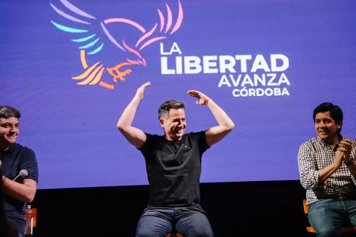 Tres hombres sentados en un escenario frente a una pantalla con el logo de un águila y el texto La Libertad Avanza Córdoba mientras uno de ellos sonríe y levanta los brazos Tres hombres sentados en un escenario frente a una pantalla con el logo de un águila y el texto La Libertad Avanza Córdoba mientras uno de ellos sonríe y levanta los brazos