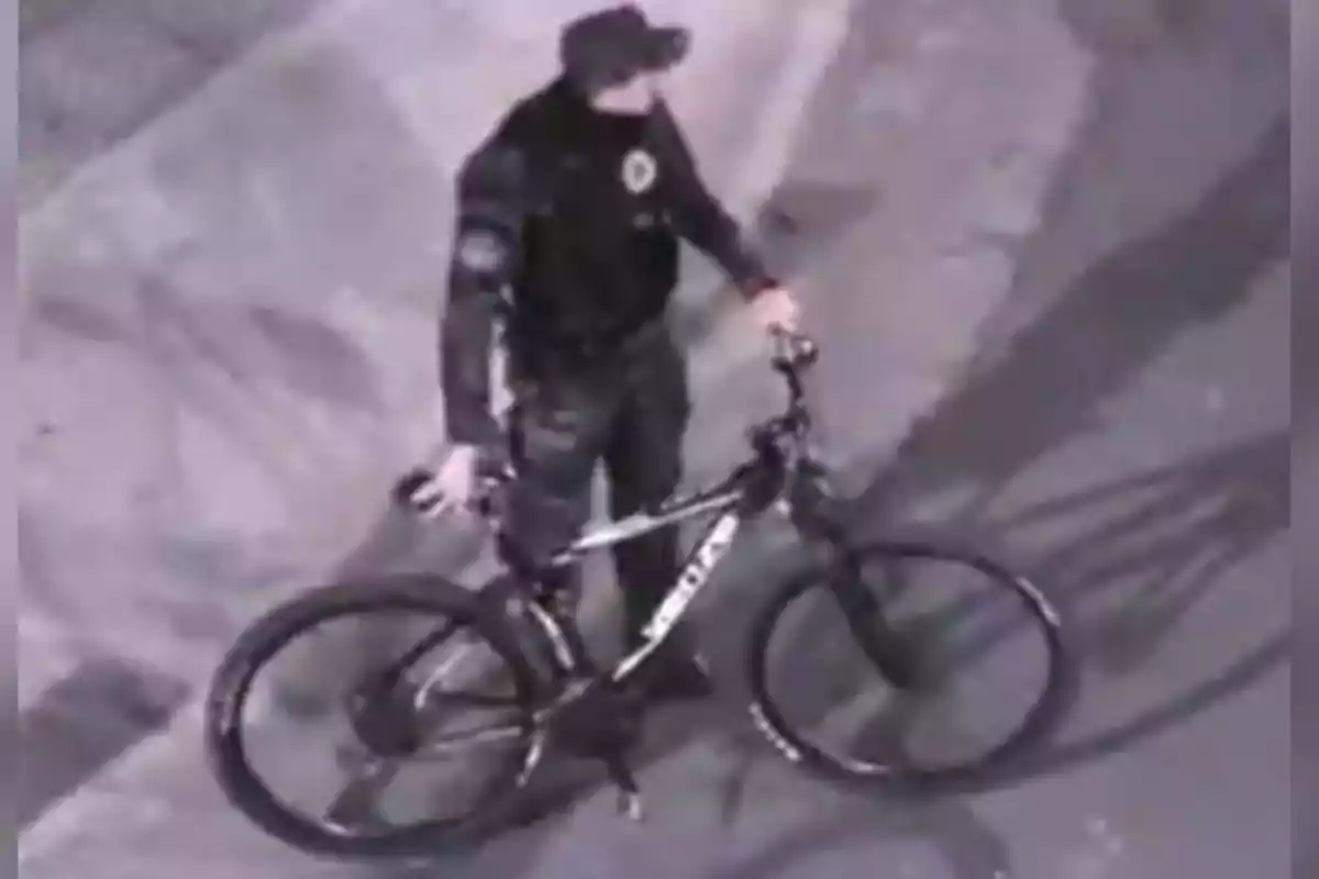 Person in a dark uniform and cap standing next to a bicycle on a paved street Person in a dark uniform and cap standing next to a bicycle on a paved street