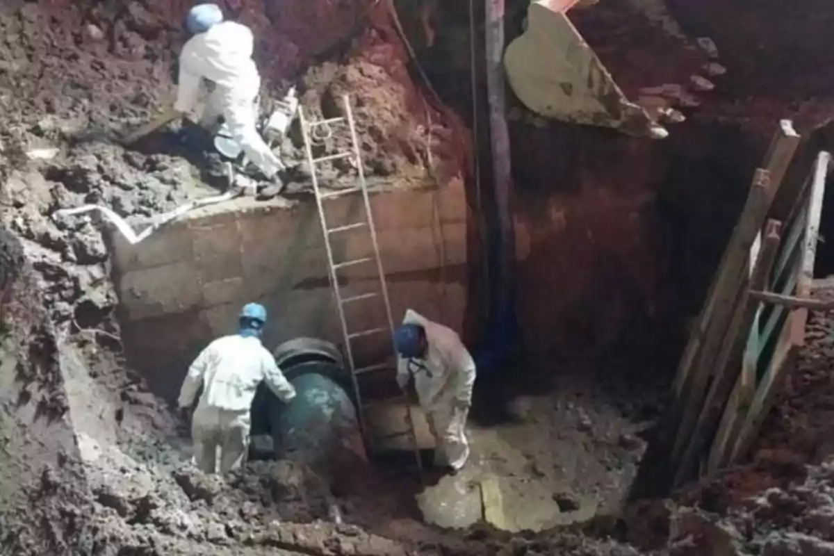 Three workers wearing helmets and protective gear are performing repair work on an underground pipe inside a trench surrounded by soil and machinery. Three workers wearing helmets and protective gear are performing repair work on an underground pipe inside a trench surrounded by soil and machinery.