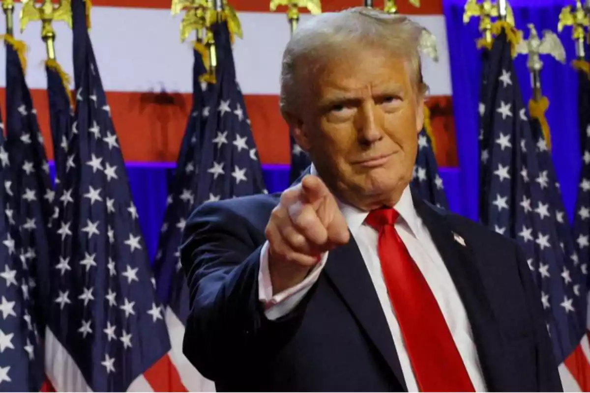 Hombre de cabello rubio con traje oscuro y corbata roja señalando al frente con varias banderas de Estados Unidos al fondo
