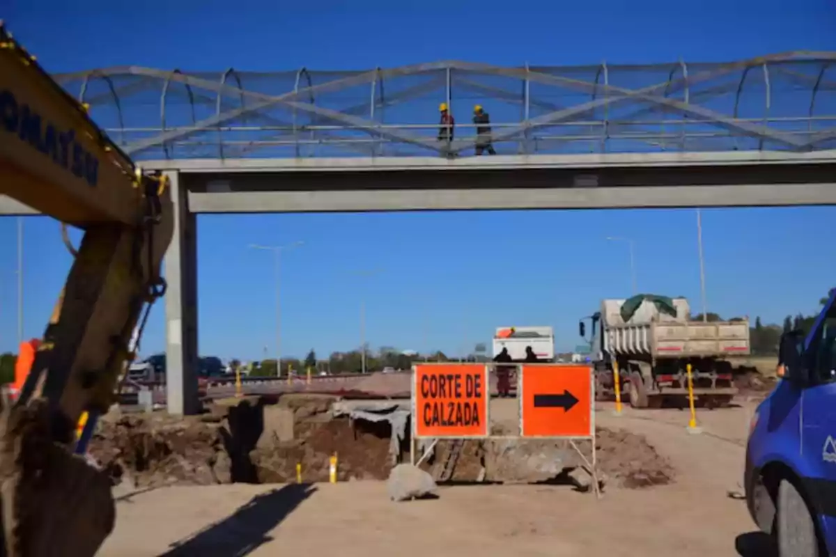 Roadworks with an elevated pedestrian bridge, workers wearing helmets, and an orange sign indicating a road closure with a black arrow. Roadworks with an elevated pedestrian bridge, workers wearing helmets, and an orange sign indicating a road closure with a black arrow.