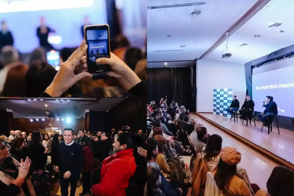 Personas asistiendo a una conferencia en un auditorio, algunas aplaudiendo y otras tomando fotos con el celular mientras tres ponentes hablan en el escenario