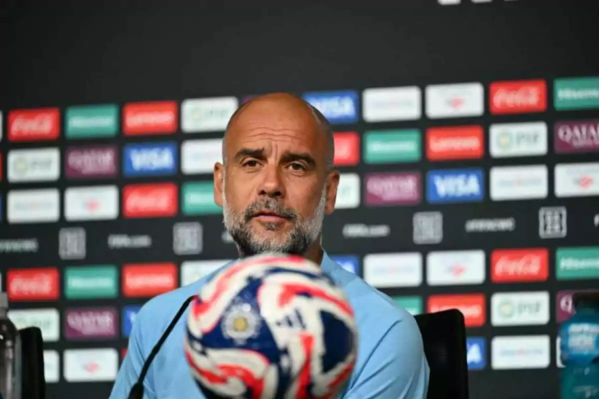Hombre calvo con barba sentado en una conferencia de prensa con un balón de fútbol en primer plano y un fondo de logotipos de patrocinadores.