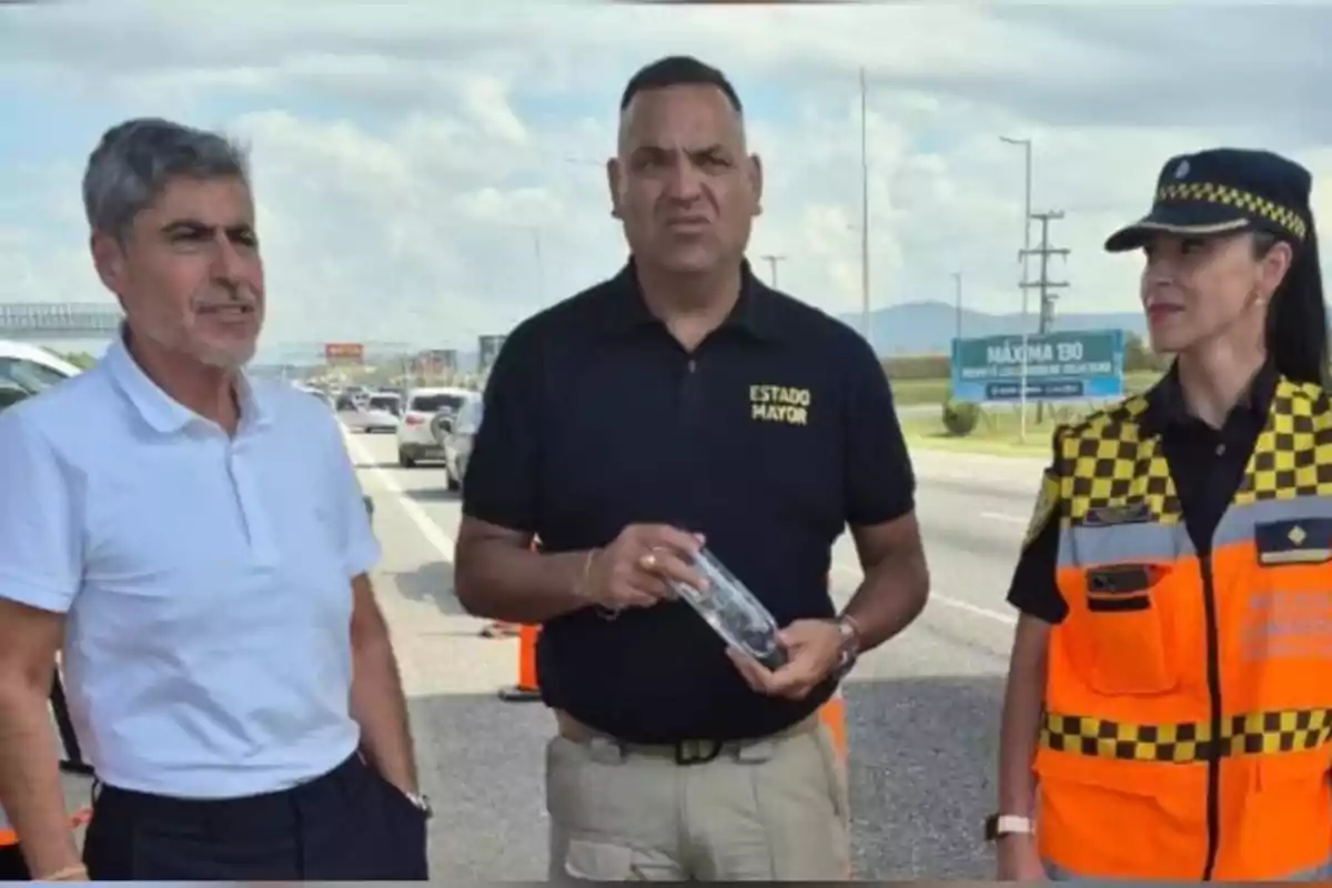 Tres personas de pie junto a una carretera, una de ellas lleva uniforme de policía vial y otra viste una camiseta con la leyenda Estado Mayor, mientras que la tercera persona lleva una camisa blanca