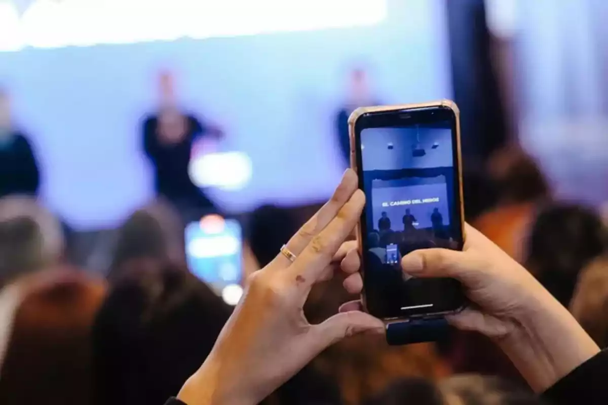 Persona sosteniendo un teléfono móvil y grabando un evento frente a una audiencia con un escenario al fondo Persona sosteniendo un teléfono móvil y grabando un evento frente a una audiencia con un escenario al fondo